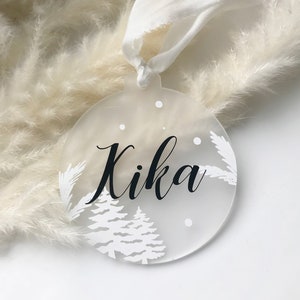 Könnte beinhalten: Eine klare, runde Weihnachtskugel mit dem Namen "Kika" in schwarzer Schrift. Der Schmuck weist weiße Winterbaum- und Schneemotive auf. Ein weißes Band ist zum Aufhängen befestigt. Der Hintergrund ist eine weiche, neutrale Farbe.