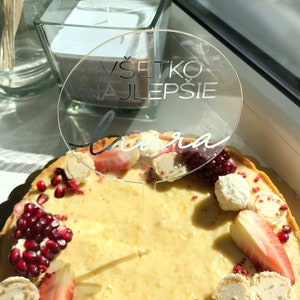 May include: A white cake decorated with red pomegranate seeds, white chocolate covered strawberries, and white chocolate candies. A clear acrylic cake topper with the text "V&scaron;etko Najlep&scaron;ie Laura" is on top of the cake.