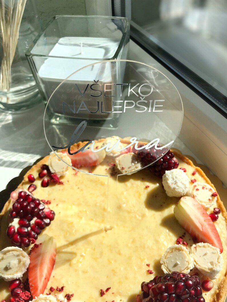 May include: A white cake decorated with red pomegranate seeds, white chocolate covered strawberries, and white chocolate candies. A clear acrylic cake topper with the text "V&scaron;etko Najlep&scaron;ie Laura" is on top of the cake.
