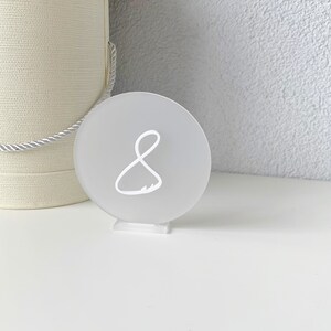 Peut inclure: Numéro de table rond en acrylique blanc givré avec le chiffre 8 écrit en blanc sur un support en acrylique transparent.