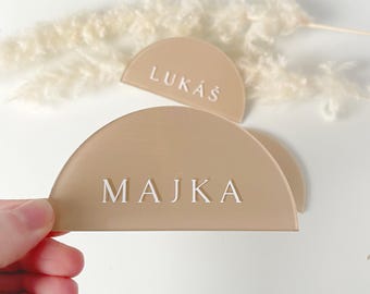 May include: Two beige acrylic half-circle place cards with white lettering. The top card reads "LUKAS" and the bottom card reads "MAJKA".