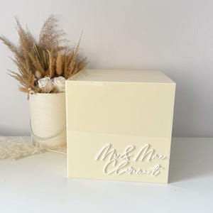 Op de afbeelding: Een lichtgele trouwkaartendoos met de woorden "Mr & Mrs Clermont" in wit script. De doos is rechthoekig en staat op een wit oppervlak. Een decoratief arrangement van gedroogde bloemen en pampasgras staat op de achtergrond.