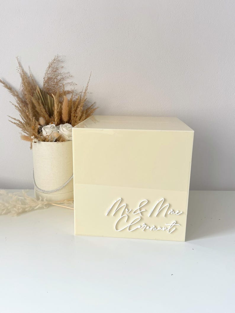 Op de afbeelding: Een lichtgele trouwkaartendoos met de woorden "Mr & Mrs Clermont" in wit script. De doos is rechthoekig en staat op een wit oppervlak. Een decoratief arrangement van gedroogde bloemen en pampasgras staat op de achtergrond.