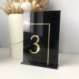 Puede incluir: Número de mesa de acrílico negro con un contorno dorado y el número "3" en dorado.