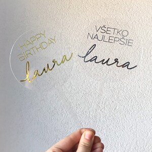 May include: A clear acrylic cake topper with gold lettering that says "Happy Birthday Laura" and black lettering that says "V&scaron;etko najlep&scaron;ie Laura".