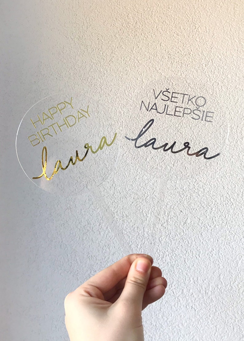 May include: A clear acrylic cake topper with gold lettering that says "Happy Birthday Laura" and black lettering that says "V&scaron;etko najlep&scaron;ie Laura".