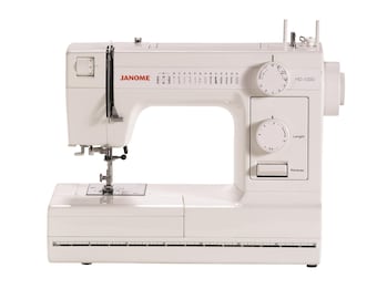 May include: A white Janome HD-1000 sewing machine featuring a stitch selection dial, a length adjustment dial, a reverse button, and a built-in ruler along the base. The machine displays various stitch patterns and the model name on the front.