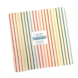 May include: A stack of fabric squares with vertical rainbow stripes. The fabric has a cream-colored background with stripes in red, orange, yellow, green, blue, and purple. A white label with text is attached to the fabric. The label reads "RUBY STAR SOCIETY warp & weft mystic fable".