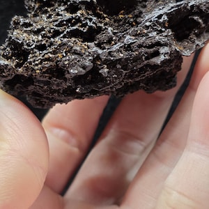 May include: A dark, porous volcanic rock specimen with a rough, uneven surface texture. The stone features deep crevices and small cavities throughout its structure, with subtle hints of metallic or rust-colored mineral deposits visible within the dark, jagged, and irregular volcanic material.