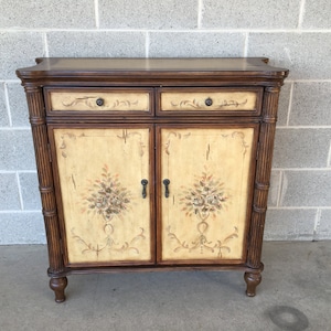 May include: A brown wooden cabinet with two drawers and two doors. The doors have a painted design of flowers and leaves on a cream background.
