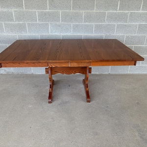 May include: A rectangular wooden dining table with a medium brown finish and visible wood grain patterns. The table features a flat top surface and is supported by a central pedestal base with two vertical legs connected by a horizontal brace.