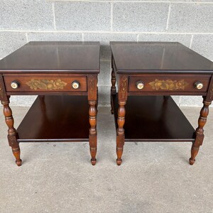 May include: Two matching dark brown wooden side tables. Each table has a drawer with floral accents and brass knobs, a lower shelf, and turned legs. The tables are approximately 24 inches wide, 24 inches deep, and 24 inches tall.