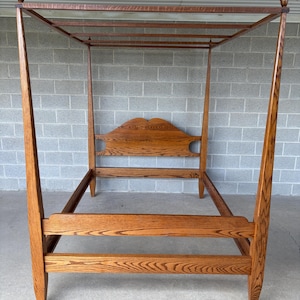 May include: A four-poster bed frame constructed from medium-toned wood with a visible grain pattern. The frame features tall, tapered corner posts topped with finials, a curved headboard, and a matching footboard with horizontal support rails.