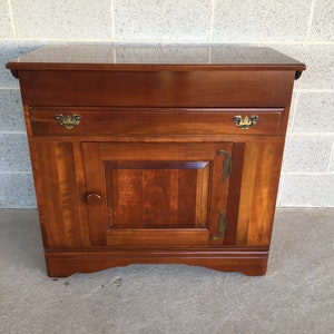 May include: A solid wood cabinet with a single drawer and a single door. The cabinet is finished in a rich brown color and has brass hardware.