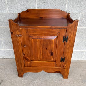 May include: A small, wooden cabinet with a warm, reddish-brown finish. The cabinet has a single door with a central panel and a small knob. The top features a curved edge, and the base has a decorative scalloped design. Black hinges are visible.