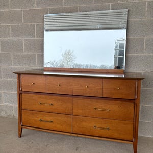 May include: A mid-century modern dresser with a matching mirror. The dresser is made of wood with six drawers and brass hardware. The mirror has a silver frame. The dresser is set against a gray brick wall.
