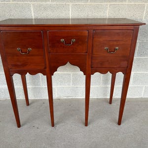 May include: A mahogany wood console table with three drawers and four legs. The table has a scalloped edge and brass hardware.