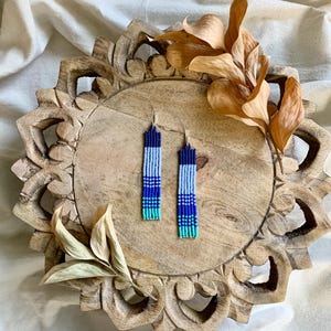 May include: A pair of long, beaded earrings with a gradient of blue, teal, and white colors. The earrings are hanging on a wooden tray with a carved design.