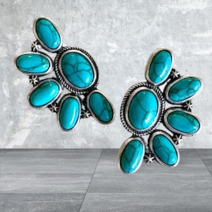 May include: A pair of silver-toned earrings featuring turquoise-colored oval stones. Each earring has a central large oval stone surrounded by smaller oval stones, creating a floral-like design. The earrings have a textured silver border.