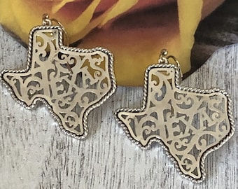 May include: A pair of gold-tone earrings shaped like the state of Texas with the word "Texas" cut out in a decorative script. The earrings have a rope-like border.