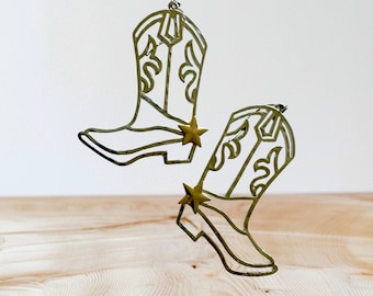 Peut inclure: Deux ornements en forme de bottes de cowboy en métal doré, chacun avec une étoile. Les bottes présentent des découpes décoratives et sont suspendues devant un fond blanc et une surface en bois. Hauteur des bottes : environ 15 cm.