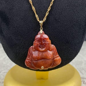 May include: A gold necklace with a pendant featuring a carved, smiling Buddha figure. The pendant is a reddish-brown color and has a smooth, polished finish. The necklace is displayed on a black velvet background.