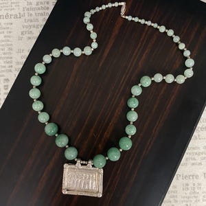 May include: A necklace with light green, round beads and smaller silver beads. It features a silver rectangular pendant with an engraved design. The necklace is displayed on a dark wooden surface.