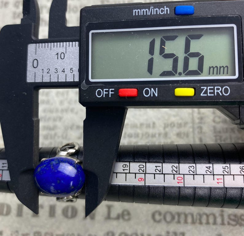 Op de afbeelding: Een zilveren ring met een grote, ovale, donkerblauwe lapis lazuli-steen. De ring wordt gemeten met een schuifmaat, die 15,6 mm aangeeft.