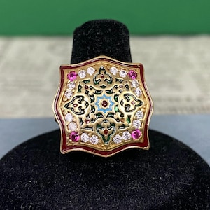Peut inclure: Une bague dorée avec un motif carré orné. La bague présente une bordure rouge, un motif central avec des accents bleus, verts et dorés, et est incrustée de pierres précieuses blanches et roses. La bague est présentée sur un support noir.