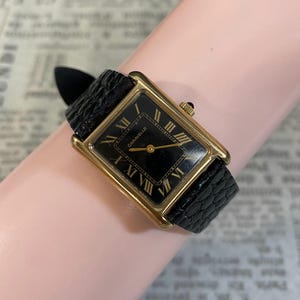 May include: A rectangular wristwatch with a gold-tone case and a black dial featuring gold Roman numeral hour markers. The watch has a black textured leather strap and the brand name Caravelle is printed in gold on the center of the dial.