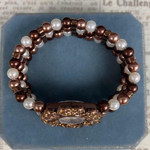 May include: A brown and white beaded bracelet with a floral watch face. The watch face is surrounded by small, sparkling crystals. The bracelet is made of brown and white beads and is set against a blue background.