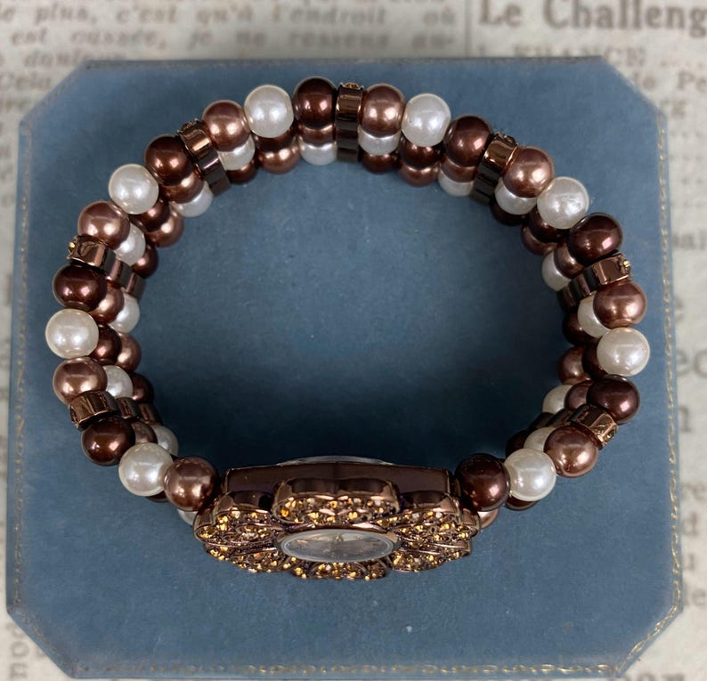 May include: A brown and white beaded bracelet with a floral watch face. The watch face is surrounded by small, sparkling crystals. The bracelet is made of brown and white beads and is set against a blue background.