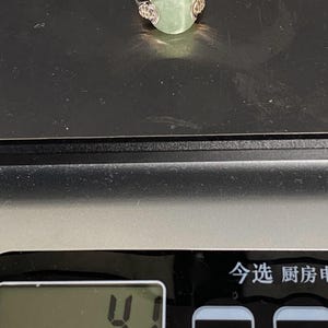May include: A silver ring with a light green, rounded gemstone. The ring is set on a digital scale, with a display showing the number 4.1. The ring's design includes decorative elements on either side of the gemstone.