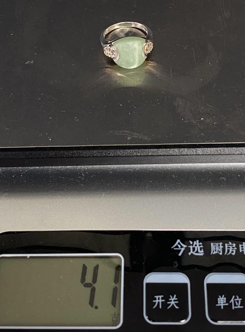 May include: A silver ring with a light green, rounded gemstone. The ring is set on a digital scale, with a display showing the number 4.1. The ring's design includes decorative elements on either side of the gemstone.