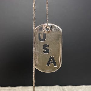 May include: A silver-toned metal dog tag pendant hanging from a thin matching chain. The rectangular tag features the letters U, S, and A cut out vertically in a stacked arrangement, creating a negative space design against the metallic surface.