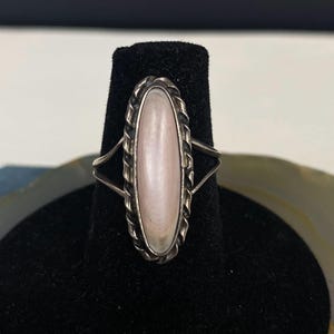 May include: A silver ring featuring an oval, pale pink stone. The stone is framed by a twisted rope-style border. The ring has a split shank design, with two bands extending from the sides of the stone.