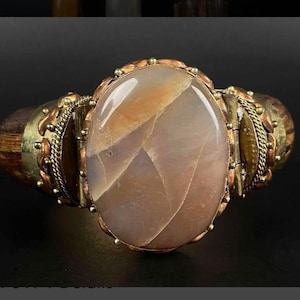 May include: A gold and copper bracelet with a large oval-shaped agate stone in the center. The stone has a light brown and white pattern. The bracelet is decorated with small beads and a chain.