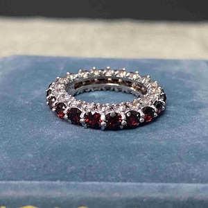 Op de afbeelding: Een zilveren eternity-ring met een doorlopende rij rondgeslepen, dieprode granaat-edelstenen in een klauwzetting. De ring is vervaardigd uit gepolijst metaal en heeft een cirkelvormig ontwerp met een gelijkmatige plaatsing van de stenen.