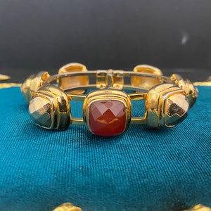 May include: A gold bangle bracelet with five square-shaped red gemstones. The bracelet is open and has a hinge closure.