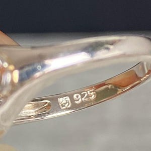 May include: Close-up of a silver ring with a polished finish. The ring is stamped with the number "925", indicating the silver content. The ring's band is smooth and reflective, with a slight curve.