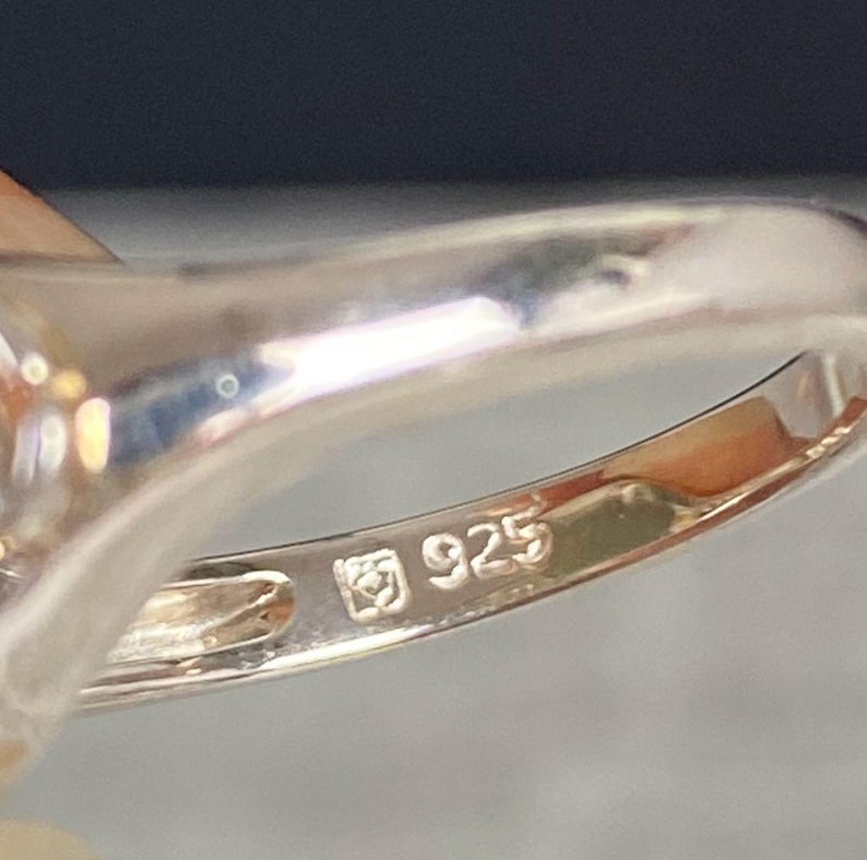 May include: Close-up of a silver ring with a polished finish. The ring is stamped with the number "925", indicating the silver content. The ring's band is smooth and reflective, with a slight curve.