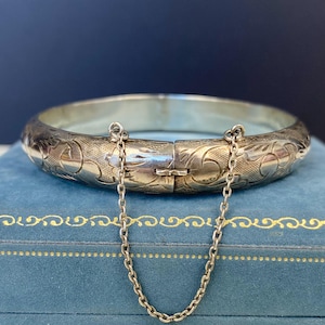 May include: Silver hinged bangle bracelet with detailed floral and leaf engravings. The bracelet features a secure clasp and a safety chain. The jewellery is displayed on a blue surface.