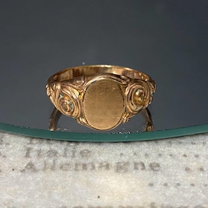 Op de afbeelding: Een goudkleurige ring met een centrale platte plaat in de vorm van een ovaal. De band is aan beide zijden van het centrale ovaal versierd met verhoogde, wervelende krulpatronen. Het metaal heeft een gepolijste afwerking met zichtbare lichtreflecties.