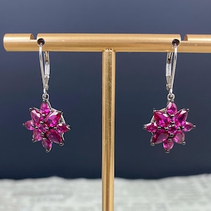 May include: A pair of silver drop earrings. Each earring showcases a cluster of bright fuchsia gemstones, arranged in a floral pattern. The teardrop-shaped gemstones are set in a silver frame, creating a sparkling, eye-catching accessory. The earrings are displayed on a gold stand.