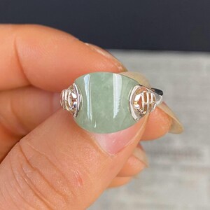 May include: A silver ring featuring a large, oval-shaped, pale green jade stone. The ring has intricate silver filigree detailing on the sides of the stone. The jade has a smooth, polished surface. The ring is a piece of jewelry.