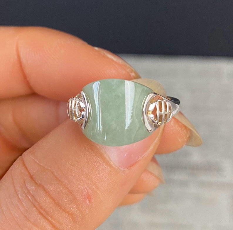 May include: A silver ring featuring a large, oval-shaped, pale green jade stone. The ring has intricate silver filigree detailing on the sides of the stone. The jade has a smooth, polished surface. The ring is a piece of jewelry.
