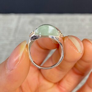 May include: A silver ring featuring a large, oval, pale green gemstone. The gemstone is set in a silver band with small, decorative accents on either side. The ring is held in a hand, showcasing its design and color.