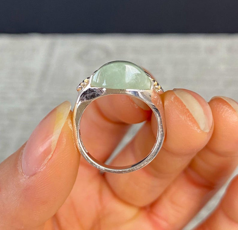 May include: A silver ring featuring a large, oval, pale green gemstone. The gemstone is set in a silver band with small, decorative accents on either side. The ring is held in a hand, showcasing its design and color.