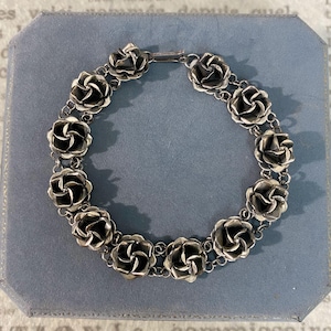 Peut inclure: Un bracelet en métal argenté composé de onze maillons individuels en forme de rose reliés par de petits anneaux circulaires. Les maillons floraux présentent une finition sombre et oxydée à l'intérieur des pétales et polie sur les bords.