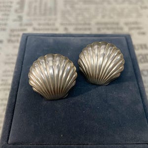 Op de afbeelding: Een paar zilverkleurige oorbellen in de vorm van sint-jakobsschelpen. Elke oorbel heeft een waaiervormig ontwerp met verhoogde verticale ribbels die vanuit de basis naar de geschulpte rand lopen, wat zorgt voor een gestructureerd oppervlak.